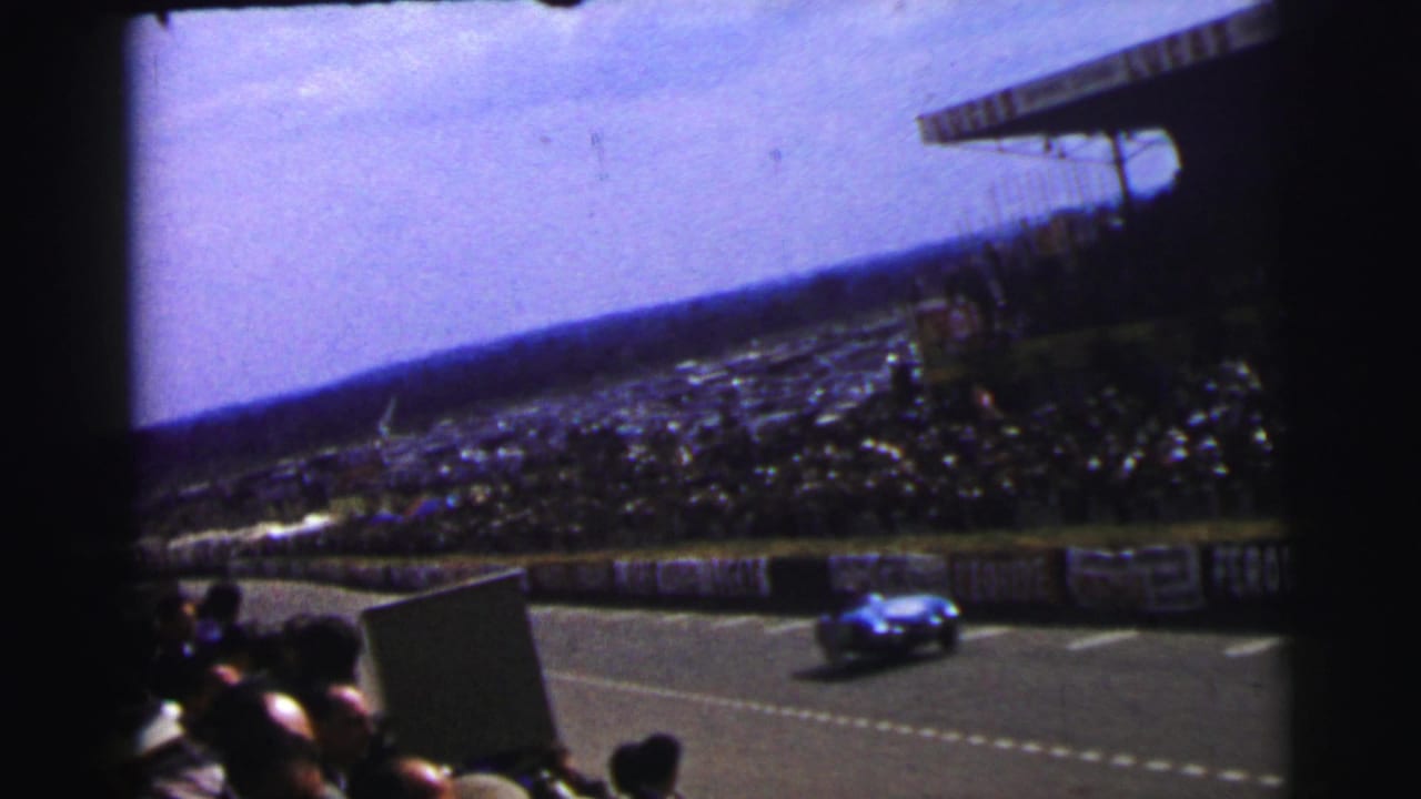Home-movie frame from Le Mans, France, with a blue racing car passing advertising barriers before a large spectator crowd.