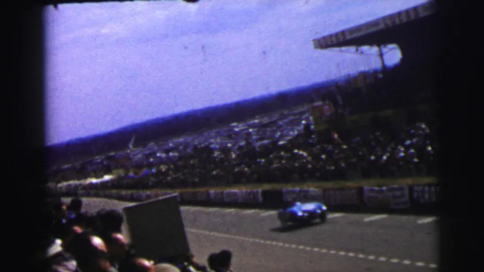 Home-movie frame from Le Mans, France, with a blue racing car passing advertising barriers before a large spectator crowd.