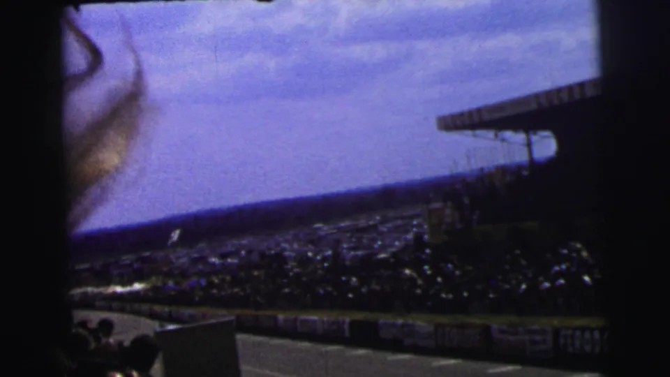 Visitor-shot 8mm view at Le Mans with the track, crowd, roof canopy, and a strand of hair briefly entering the frame.