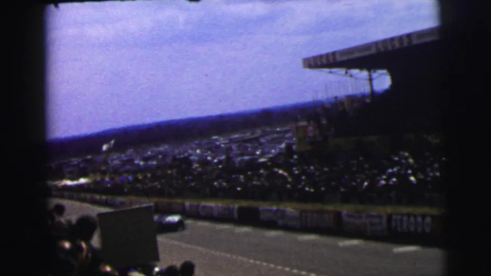 1954 Le Mans crowd scene from the stands, with advertising barriers, a packed spectator area, and a blue car near the track edge.