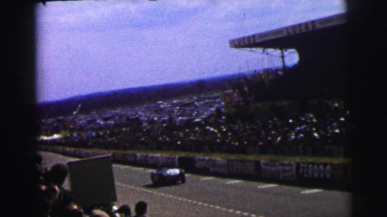 1954 Le Mans spectator view of a race car on the circuit below crowded stands and trackside banners.