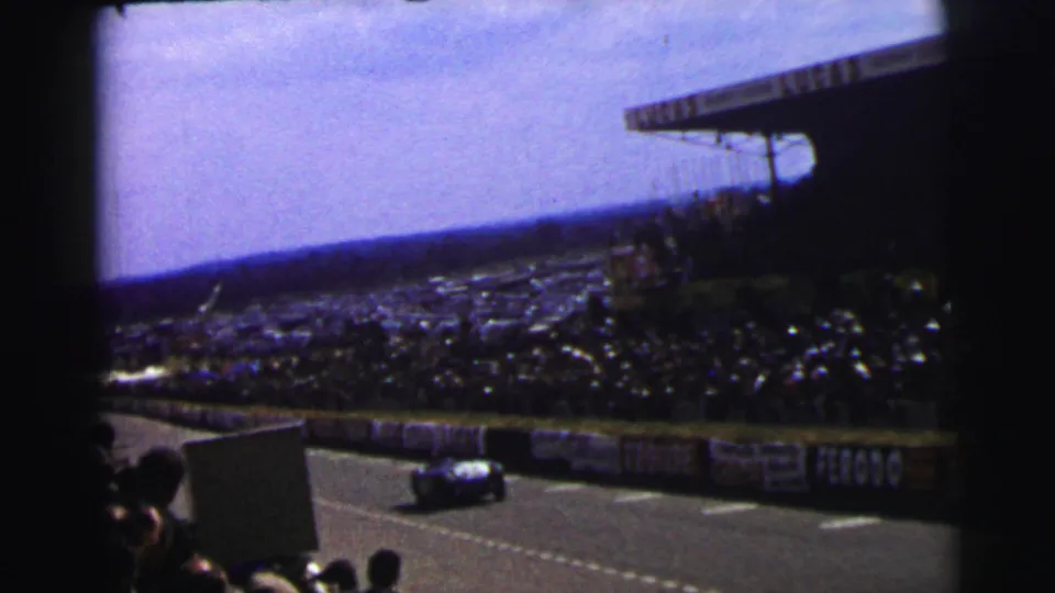 1954 Le Mans spectator view of a race car on the circuit below crowded stands and trackside banners.