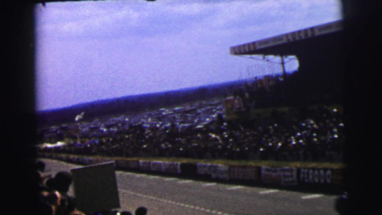Spectator-shot 1954 Le Mans view of the circuit, crowded stands, roof structure, and visible trackside advertising.