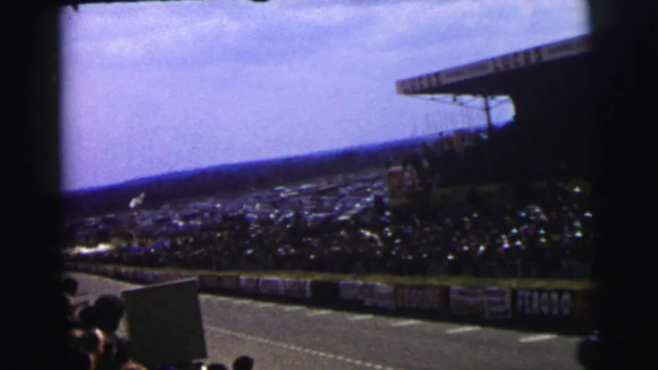 Spectator-shot 1954 Le Mans view of the circuit, crowded stands, roof structure, and visible trackside advertising.
