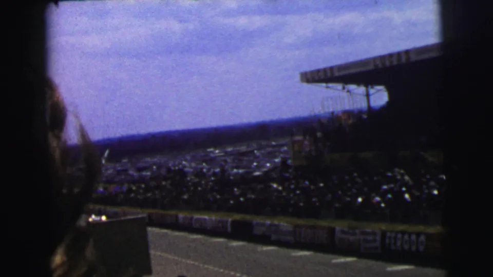 1954 Le Mans home-movie frame showing crowded stands, trackside boards, and a partial spectator silhouette near the camera.