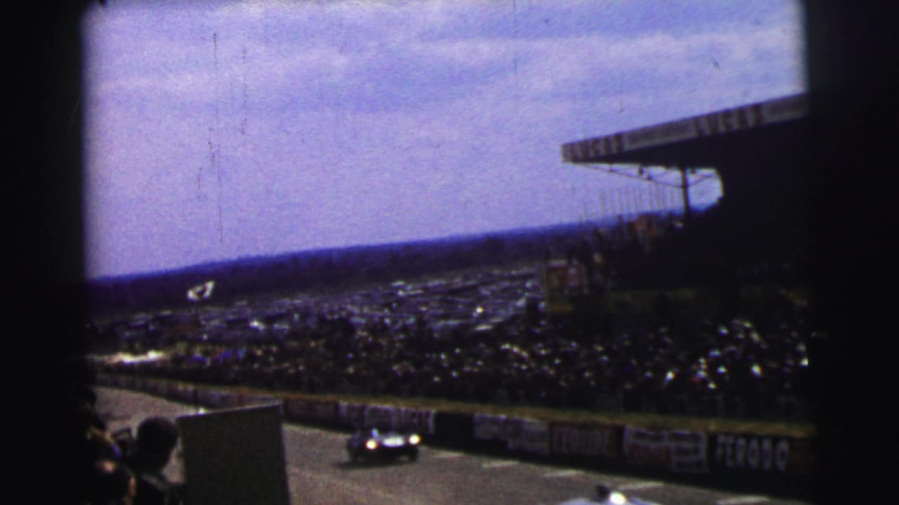 Race car passes a crowded trackside grandstand with barrier advertising visible.