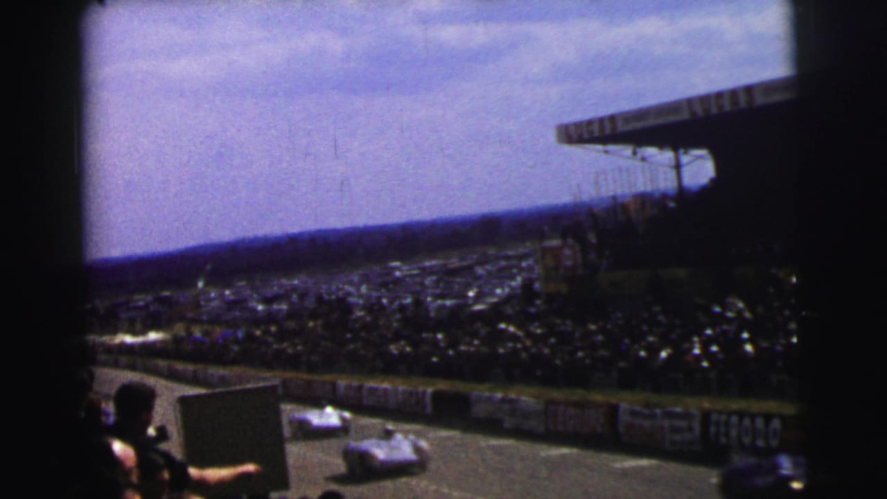 Spectator-shot 1954 Le Mans view of racing cars passing the stands, with dense crowd areas and visible barrier advertisements.