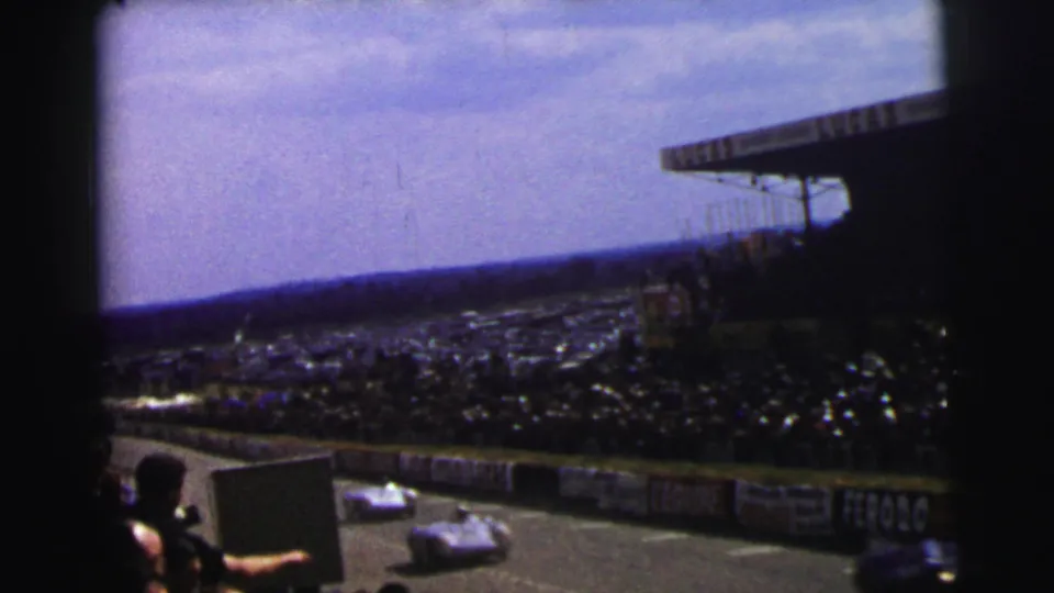 Spectator-shot 1954 Le Mans view of racing cars passing the stands, with dense crowd areas and visible barrier advertisements.