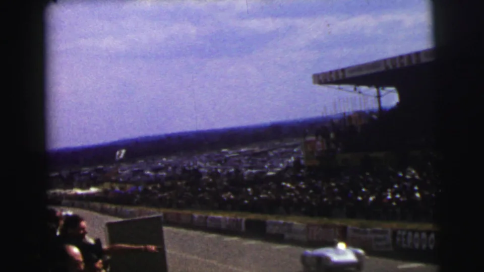 1954 Le Mans home-movie frame showing a light race car near the foreground, packed spectators, track barriers, and grandstand structure.