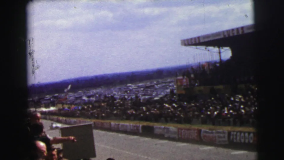 Trackside 1954 Le Mans view with a racing car entering the frame, dense spectators, advertising boards, and handheld 8mm framing.