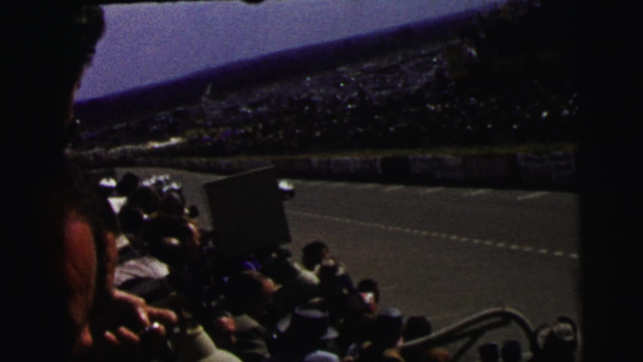 1954 Le Mans home-movie frame showing spectators in the stands as open-wheel race cars pass along the track.