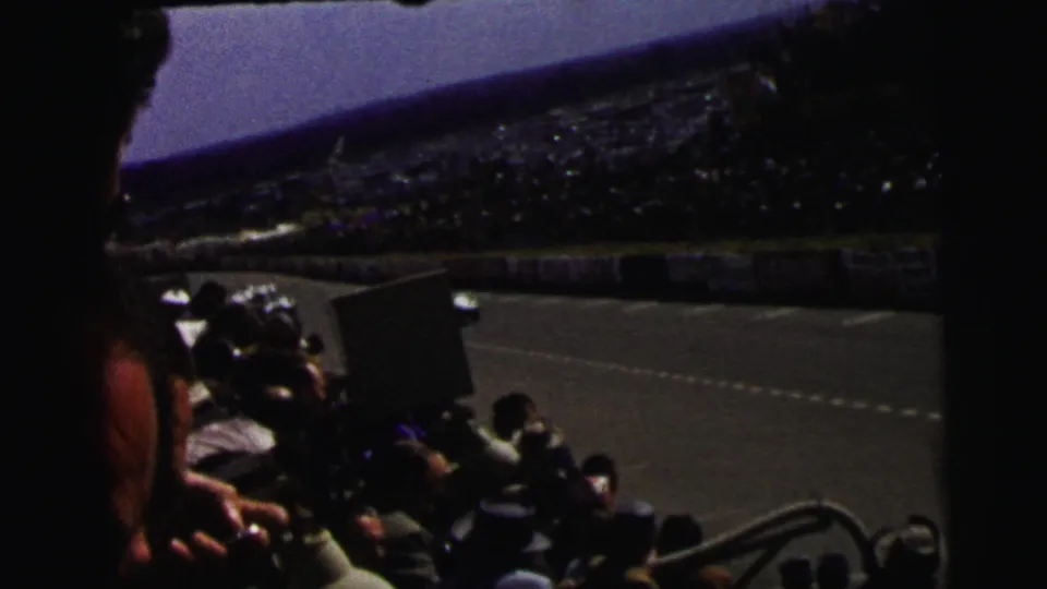 1954 Le Mans home-movie frame showing spectators in the stands as open-wheel race cars pass along the track.