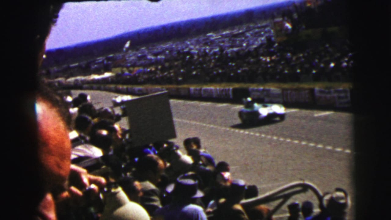 A white open-wheel car moves past the Le Mans spectator stands, with foreground viewers and trackside boards visible.