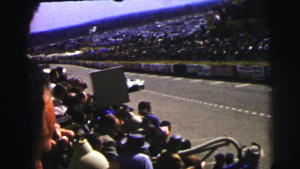 Le Mans spectator-view footage shows a race car on the track, dense grandstands, railings, and mid-century viewers in the foreground.