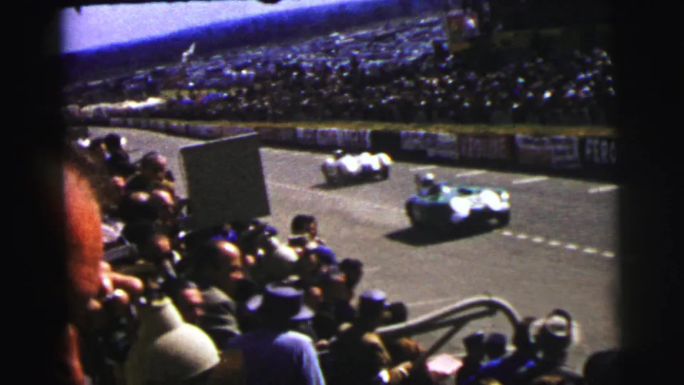 A spectator-shot 1954 Le Mans frame captures two open-wheel cars on the track before a packed grandstand.
