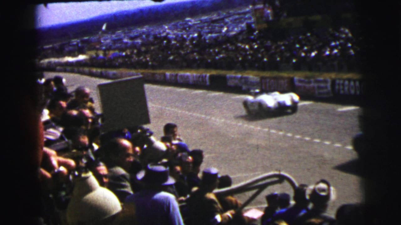 Crowded race stands beside a paved track as a pale race car passes in the distance.