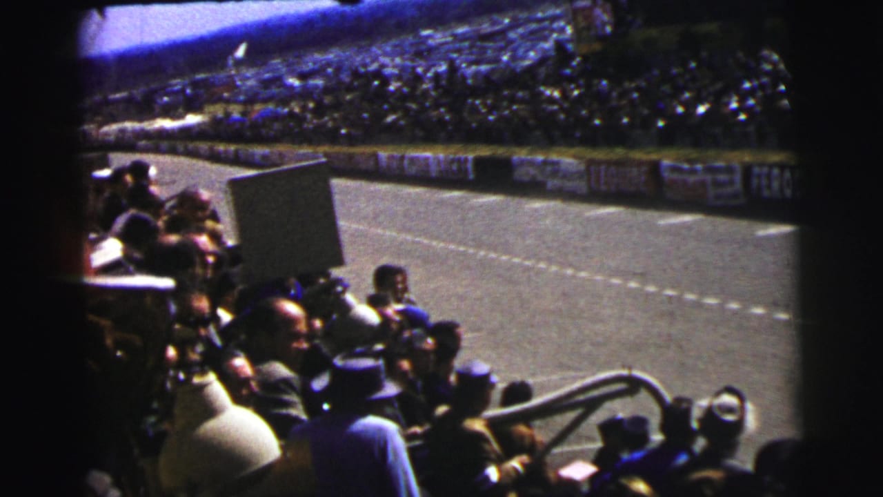 Trackside spectators at Le Mans in 1954 sit near the racing surface as sponsor boards line the opposite barrier.