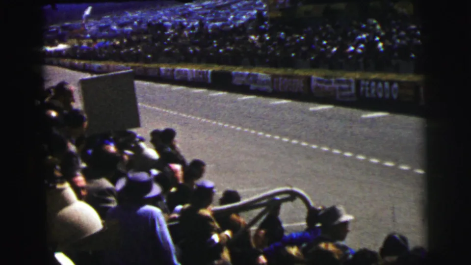 Handheld 8mm view from the Le Mans stands, looking past spectators and railing toward the track and sponsor boards.
