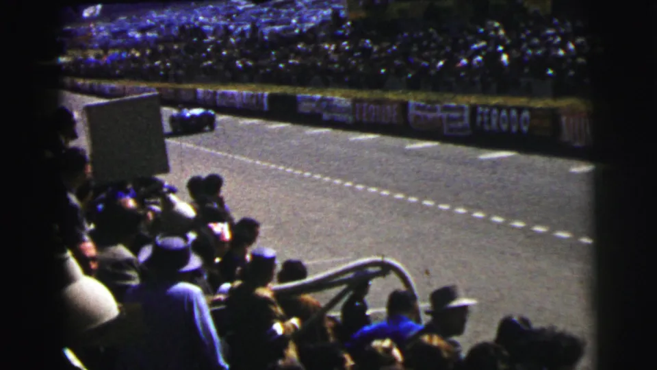 A dark race car passes along the Le Mans circuit in 1954 while spectators watch from packed trackside seating.