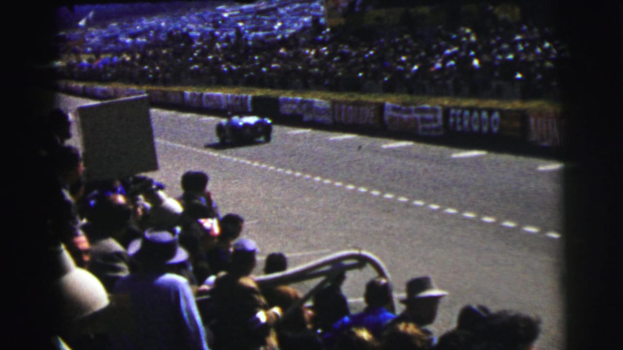 1954 Le Mans home-movie view of spectators in hats watching a dark race car move along the track past advertising boards.