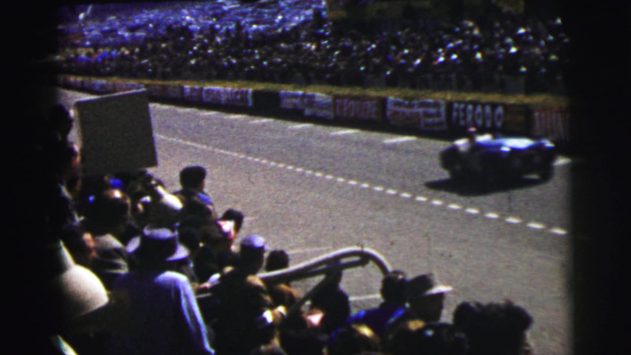 A blue race car speeds past spectators seated beside the track.
