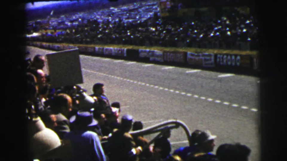 1950s Le Mans spectators sit and stand beside the road circuit, facing the track and advertising-lined barrier.