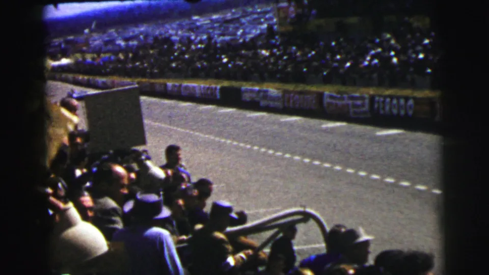 Crowded Le Mans stand seen from a home-movie angle, with spectators watching the advertising-lined racing circuit.
