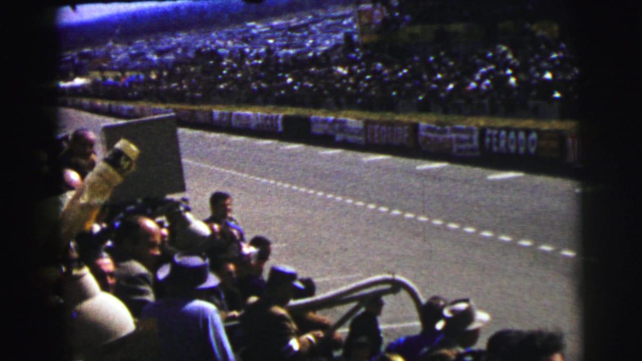 Spectators watch a race track from behind a barrier, with grandstands across the course.