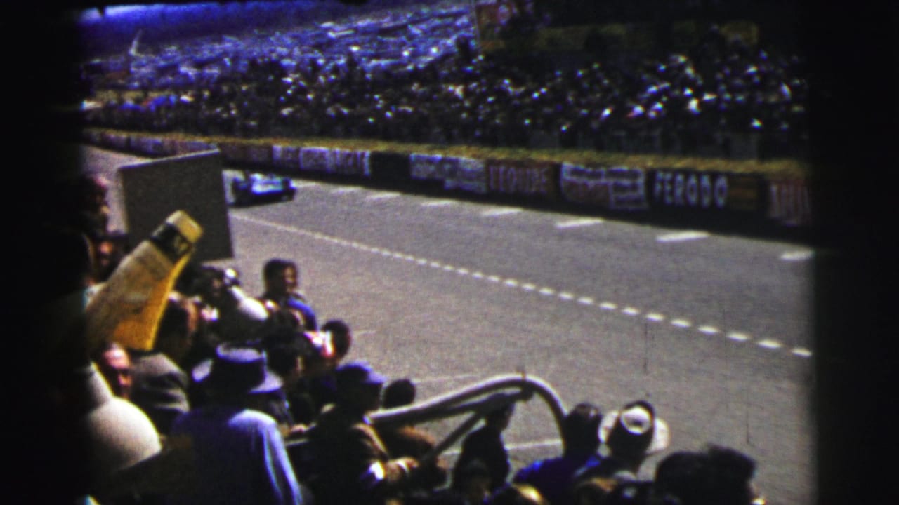 Blue racing car passes on the Le Mans circuit as spectators and trackside photographers watch from the foreground.