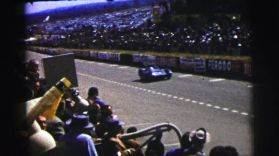 1954 Le Mans racing view with a blue car on the circuit, foreground spectators, barrier advertising, and full grandstands.