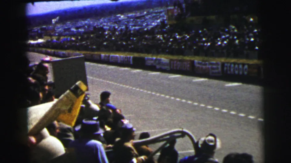 Le Mans trackside crowd seen from an 8mm home-movie angle, with empty track, barrier signs, and grandstands beyond.