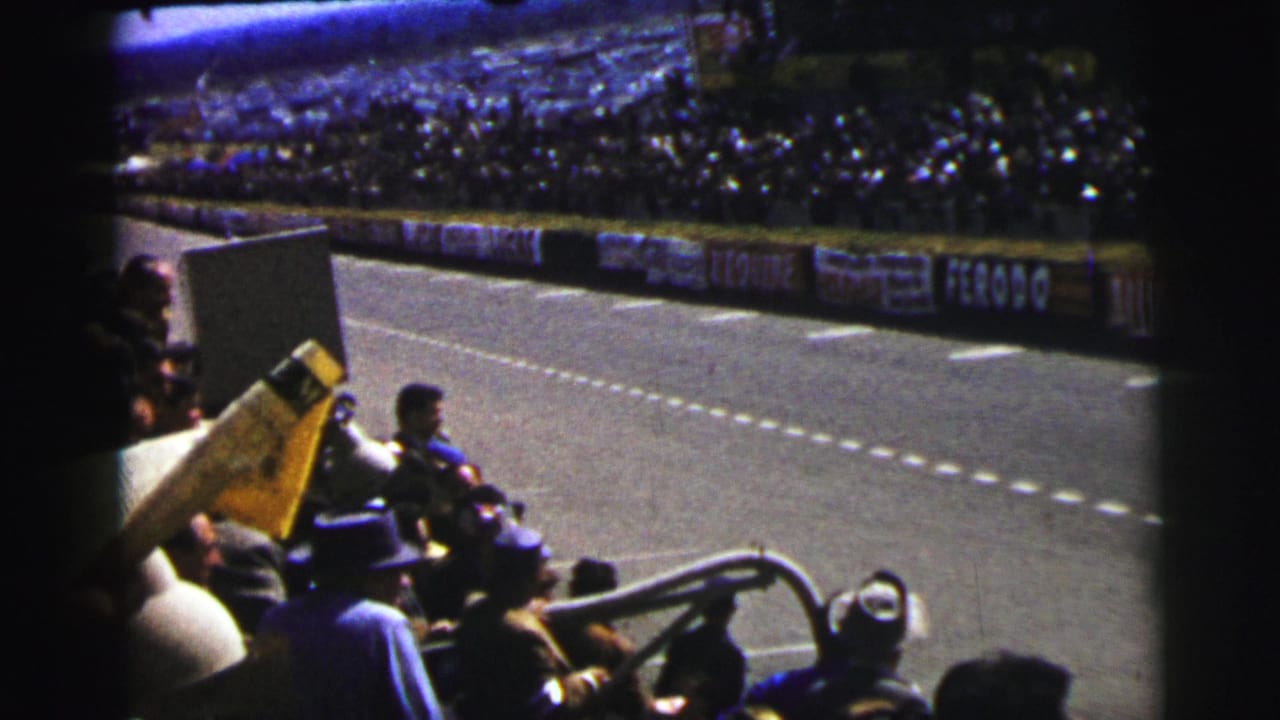 1954 Le Mans spectator view with a yellow flag in the foreground, seated fans, track lanes, and packed grandstands across the circuit.