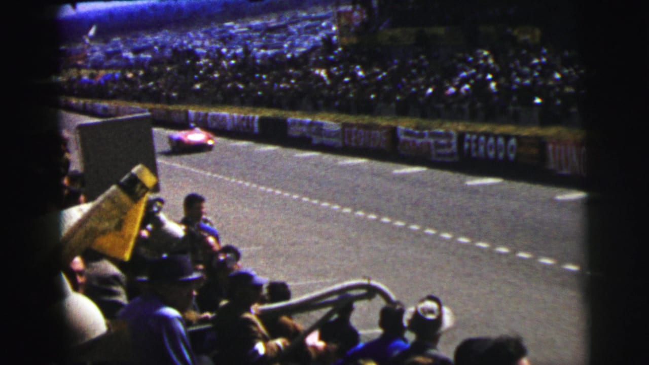 A red race car approaches along the track in front of crowded stands.