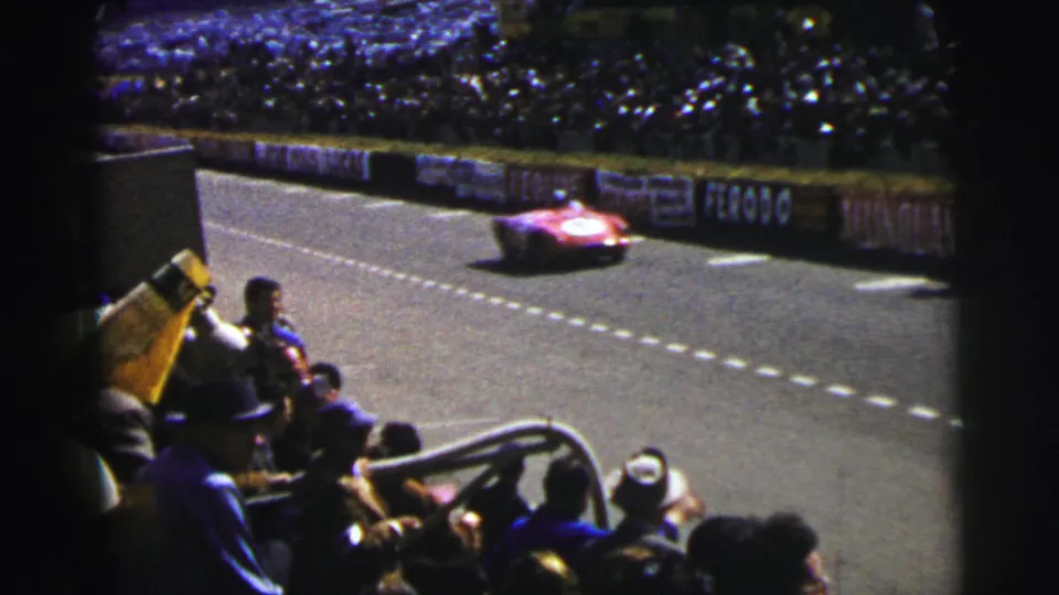 The red race car blurs past the spectator viewpoint at Le Mans, with seated fans, a yellow flag, signage, and crowd-filled stands visible.
