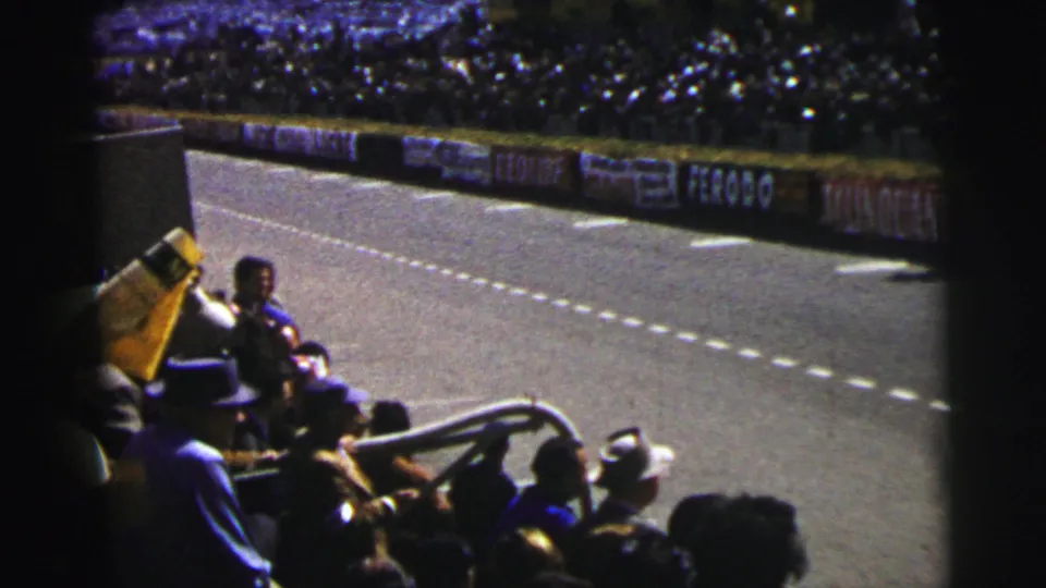 After the car passes, the frame holds on Le Mans spectators, the yellow flag, track lanes, advertising boards, and the packed grandstand beyond.