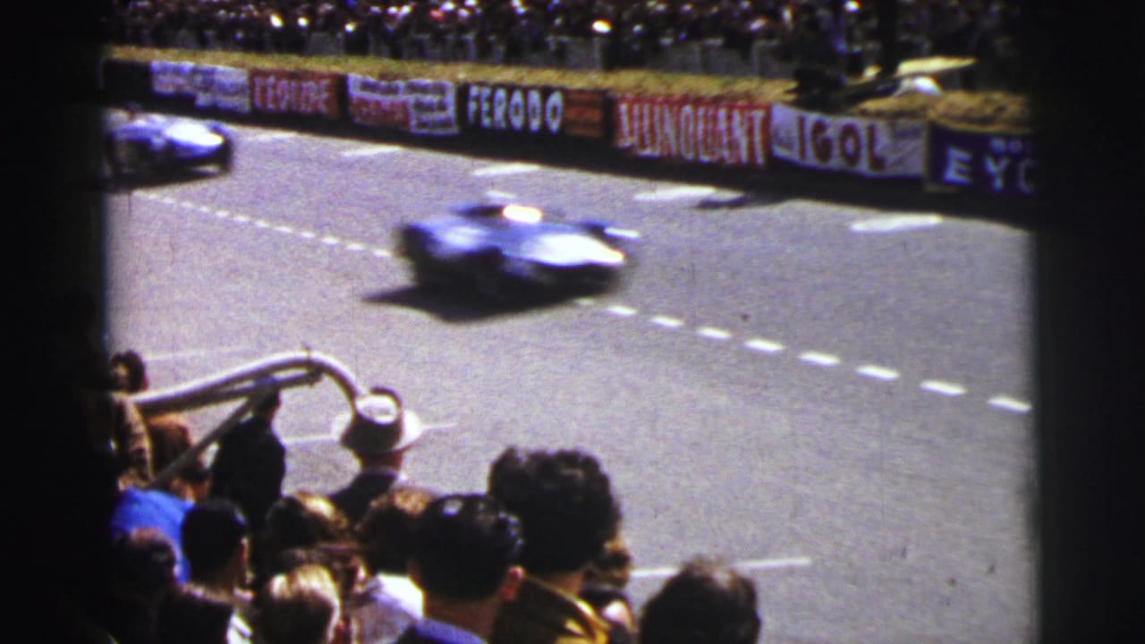 At Le Mans in 1954, a blue racing car speeds past a crowded stand with advertising banners along the circuit wall.