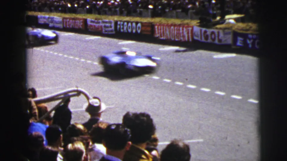 At Le Mans in 1954, a blue racing car speeds past a crowded stand with advertising banners along the circuit wall.
