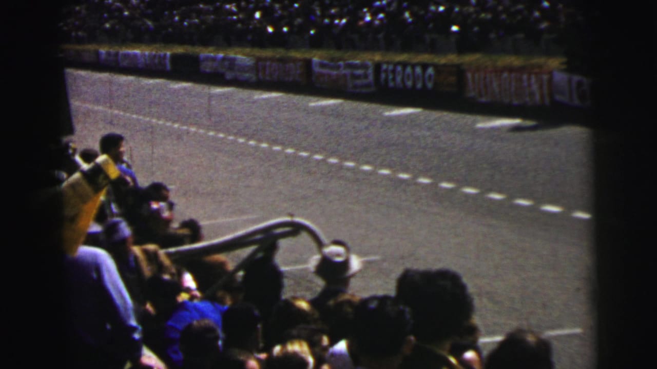 Spectators at the 1954 Le Mans race watch from bleachers as the track curves past advertising boards and lane markings.