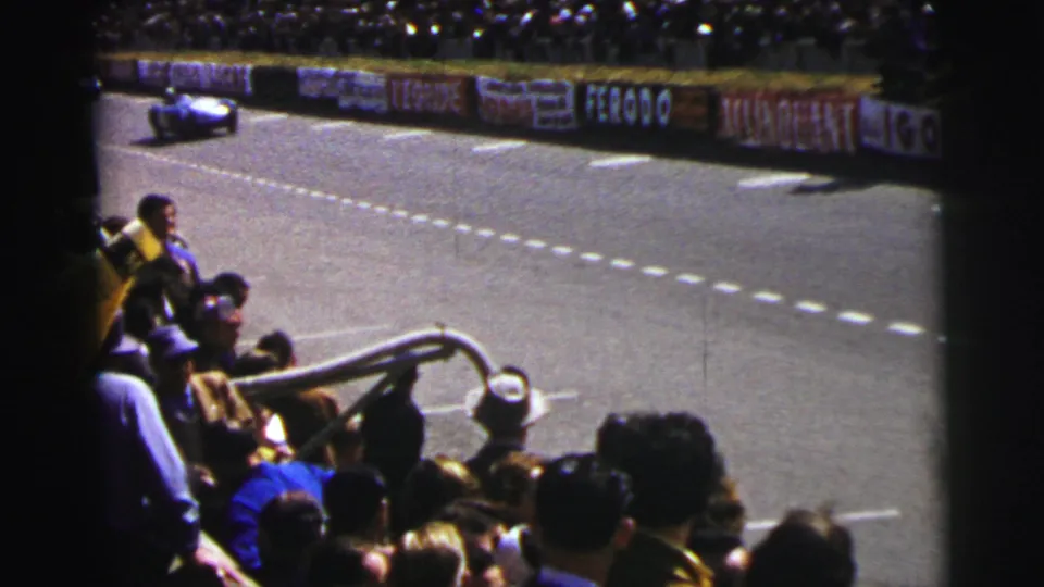 From the bleachers at 1954 Le Mans, spectators face the circuit as a race car moves out of frame near the advertising-lined track.