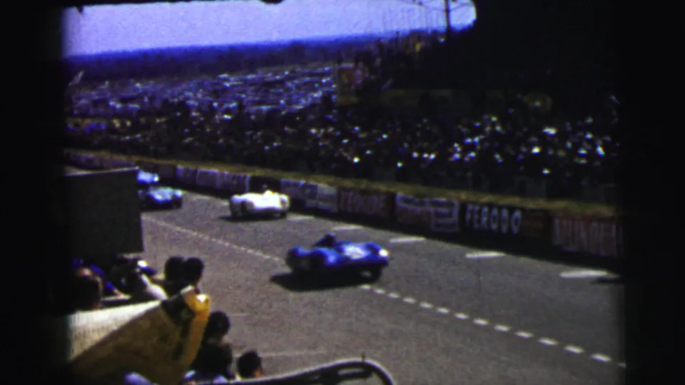 Multiple vintage racing cars pass the crowded Le Mans circuit in 1954, seen from a handheld spectator position in the stands.
