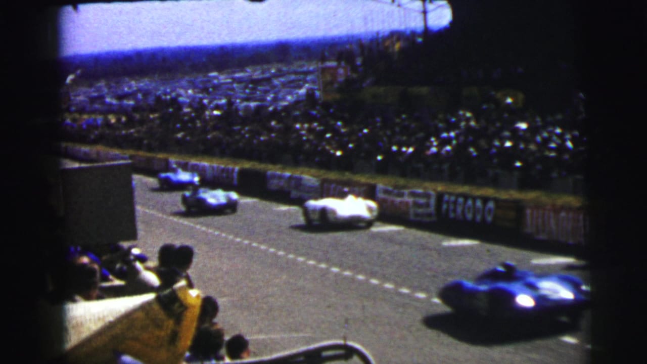 At Le Mans in 1954, blue and white race cars appear on the circuit before a dense crowd and sponsor-lined barriers.