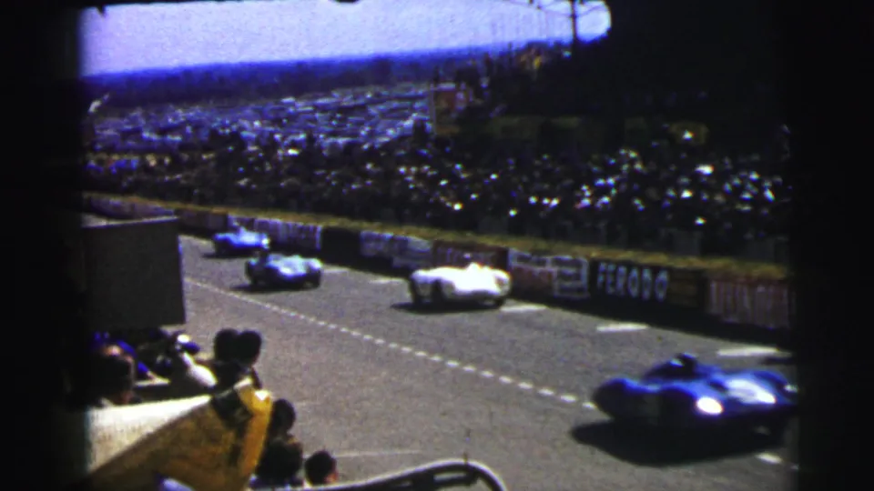 At Le Mans in 1954, blue and white race cars appear on the circuit before a dense crowd and sponsor-lined barriers.