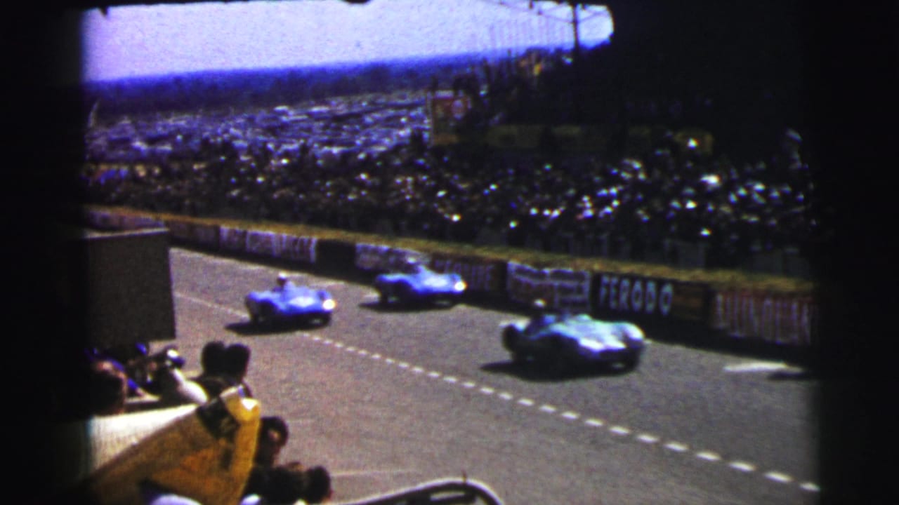 Multiple 1954 Le Mans race cars run past a packed viewing area, with spectators and advertising boards along the track edge.