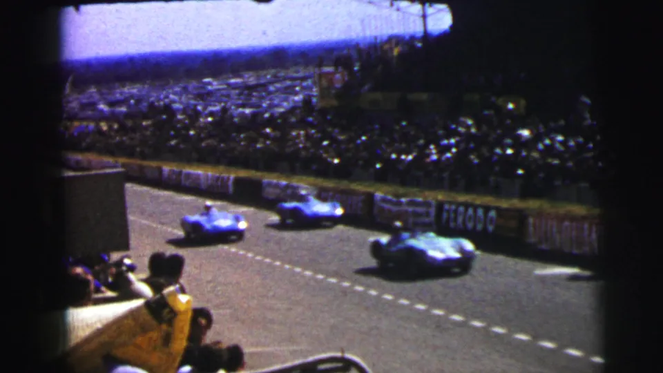 Multiple 1954 Le Mans race cars run past a packed viewing area, with spectators and advertising boards along the track edge.