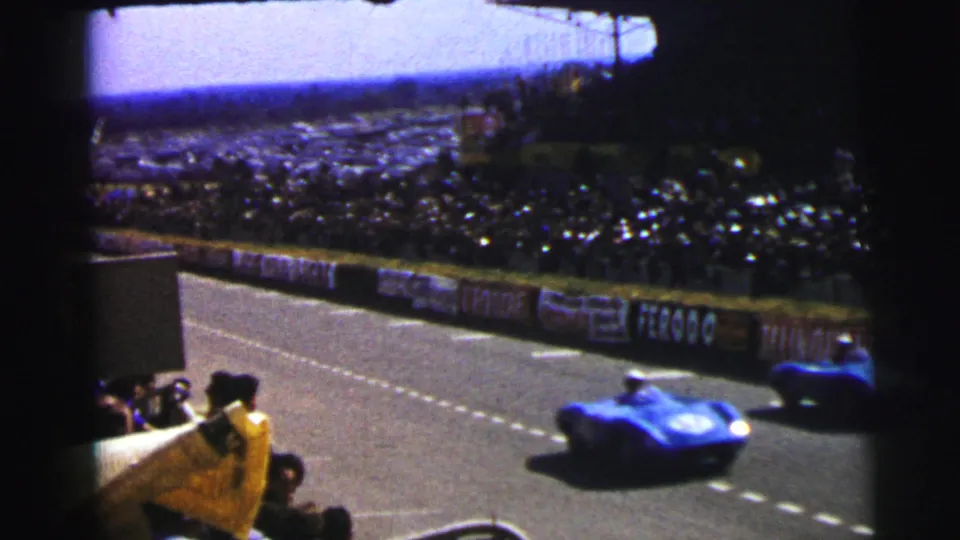 A blue race car sweeps past the spectator viewpoint at the 1954 Le Mans circuit, with another car visible farther along the barrier.