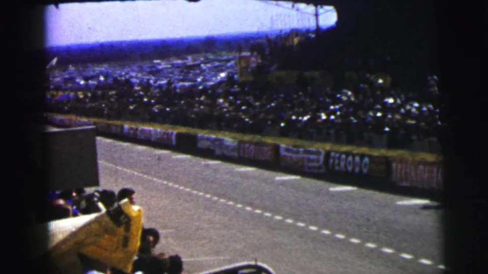 Near the end of the shot, a race car leaves the 1954 Le Mans frame while the crowd and banner-lined circuit remain visible.