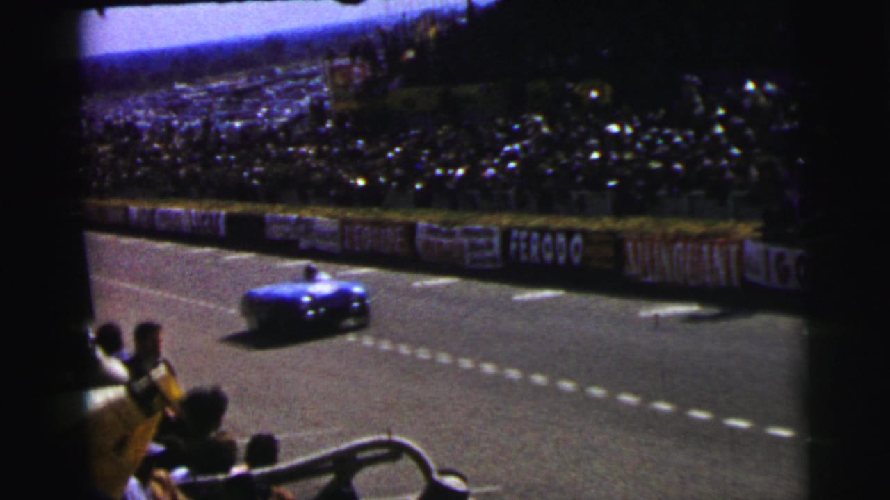 Blue race car passing spectators on a paved track with crowd and banners behind it.