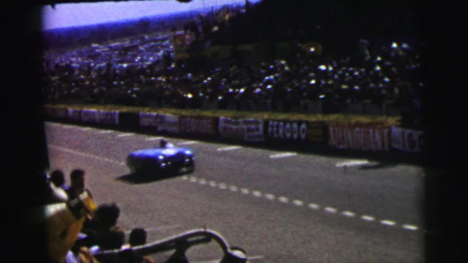 At Le Mans, France in 1954, a blue race car passes a crowd-lined track with advertising boards and spectators in the foreground.