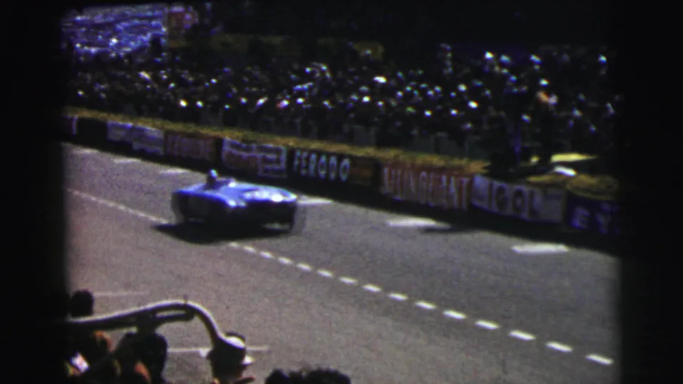 A blue race car moves along the Le Mans track in 1954 as spectators watch from behind barriers and advertising boards.