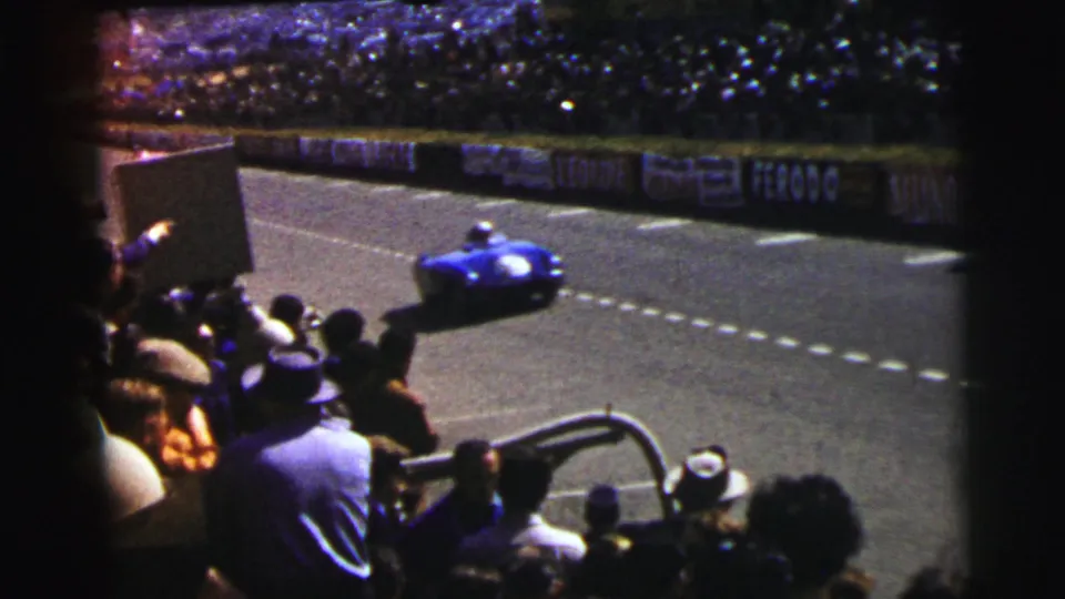 From a spectator viewpoint at Le Mans, France in 1954, a blue race car passes trackside crowds, banners, railings, and hats in the foreground.